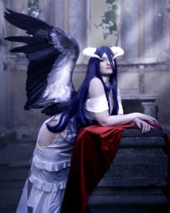 Cosplayer with long dark purple hair and black angel wings posing in a fantasy-inspired cosplay outfit with a red cloth draped beside her. Albedo cosplay