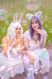 Two bunny cosplay models in white dresses sitting in a grassy field with bubbles, one with white hair and bunny ears and the other with purple hair and bunny ears