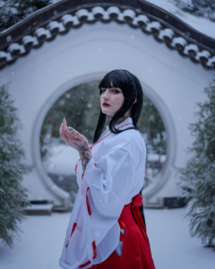 Cosplayer in a white and red anime inspired outfit standing in front of a circular stone arch in a snowy outdoor setting. Kikyo cosplay